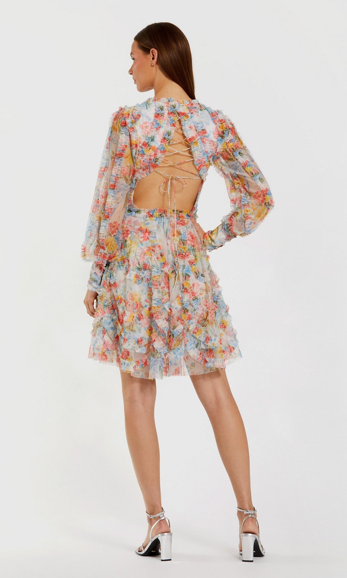 Woman models the Mac Duggal 8304 short wedding guest dress featuring a floral mesh design, long sleeves, and an open back with crisscross laces, paired with high-heeled sandals and facing away from the camera.